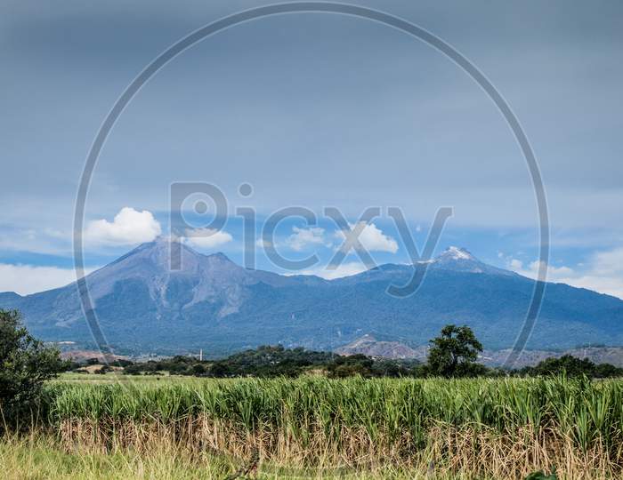 Mexican Landscape With A Sugar Cane Plantation And The Colima Volcano In The Background, Sunny Day With A Blue Sky And Abundant Clouds In The State Of Colima, Mexico
