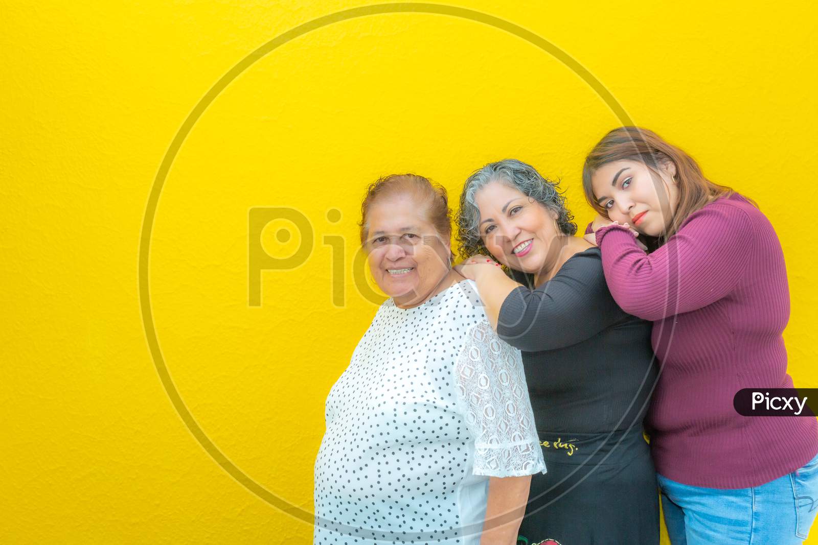 Grandmother, Daughter And Granddaughter Resting On Their Shoulders And Looking At The Camera, Three Generations Of Mexican Women Smiling In Casual Clothes In A Row On A Yellow Background