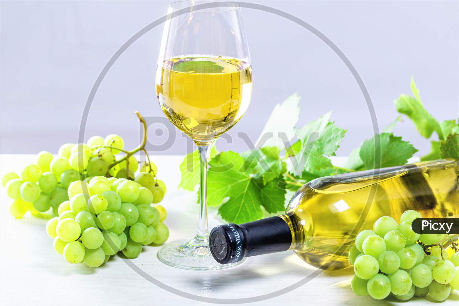 White Wine In A Glass With A Full Bottle, Grapes And Leaves On A White Wooden Background