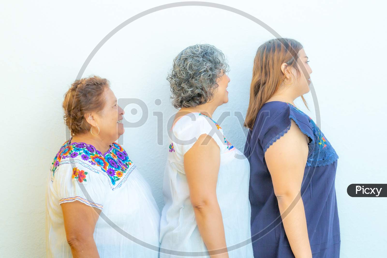 Side View Profiles Of Three Generations Of Mexican Women Smiling With Floral Printed Blouses In A Row, Grandmother, Daughter And Granddaughter On A White Background