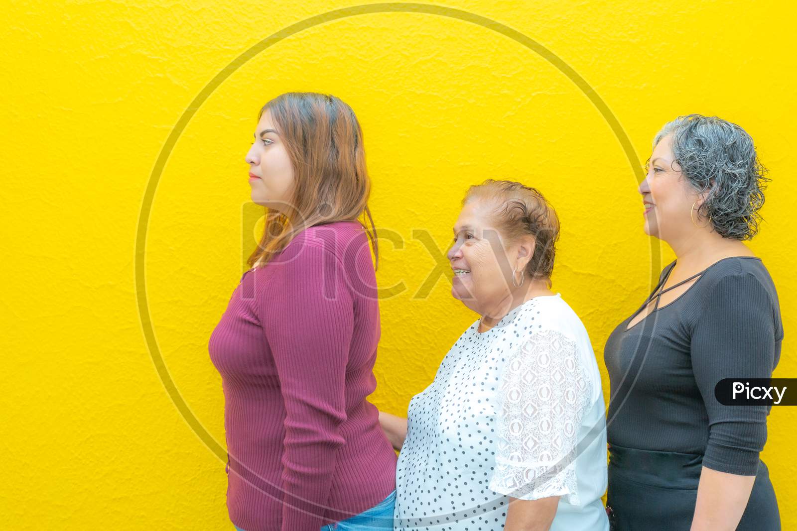 Side View Profiles Of Granddaughter Grandmother And Daughter, Three Generations Of Mexican Women Smiling With Casual Clothes In A Row On A Yellow Background