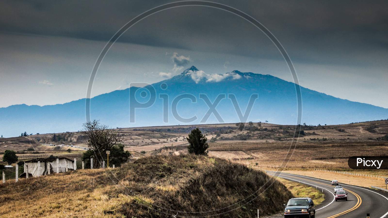 Mexican Landscape With A Curved Asphalt Highway Next To A Small Hill, Cars Driving With The Colima Volcano In The Background, Cloudy Day With Abundant Clouds In The State Of Jalisco, Mexico