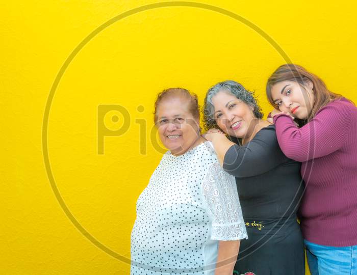Grandmother, Daughter And Granddaughter Resting On Their Shoulders And Looking At The Camera, Three Generations Of Mexican Women Smiling In Casual Clothes In A Row On A Yellow Background