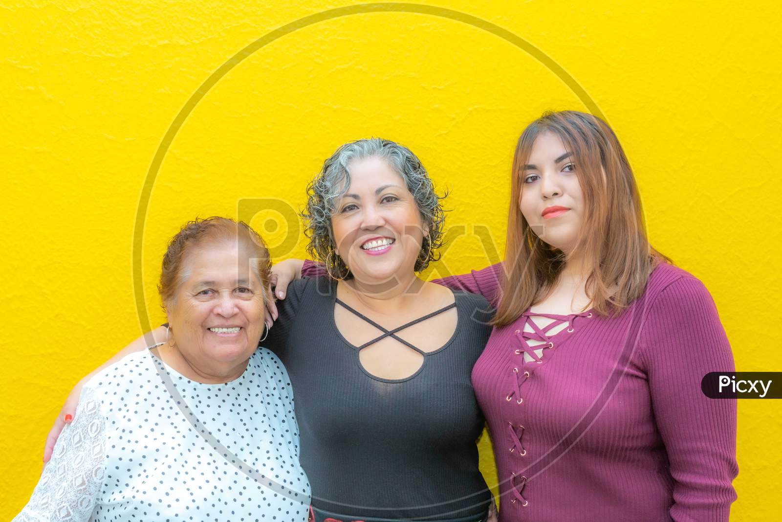 Three Generations Of Latin Mexican Women Smiling, Grandmother, Daughter And Granddaughter Wearing Casual Clothes Looking At The Camera With A Yellow Background