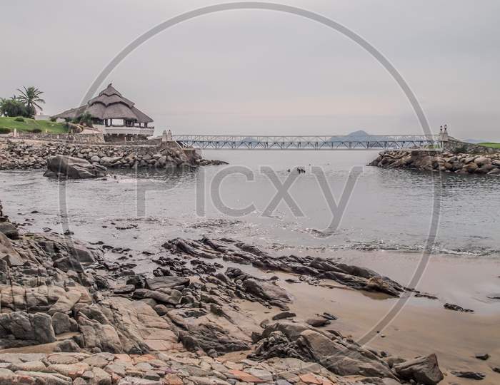 Rocky Mexican Coastal Beach With A Bay With A Palapa On The Rocks, A Small Footbridge Over The Sea In The Background, Tropical Cloudy Afternoon In Manzanillo, Colima Mexico