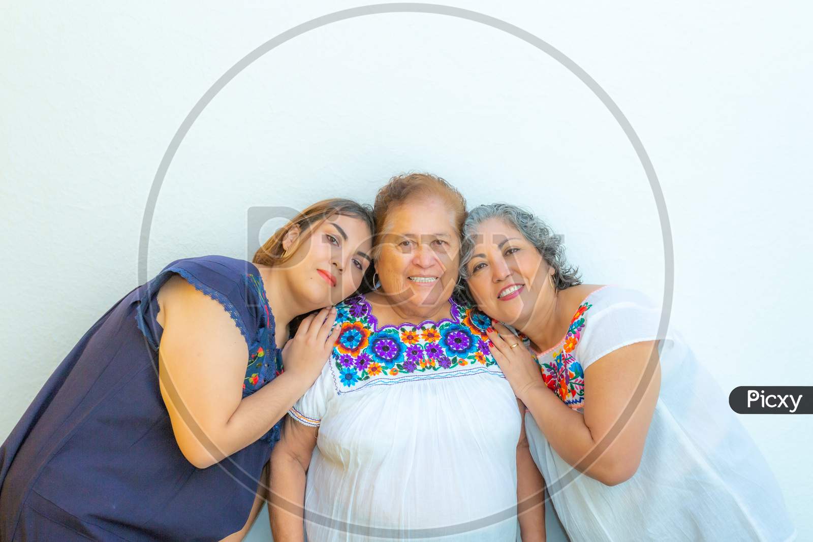 Three Generations Of Smiling Mexican Women, Mother And Daughter Leaning On Grandma, Blouses With Floral Prints On A White Background