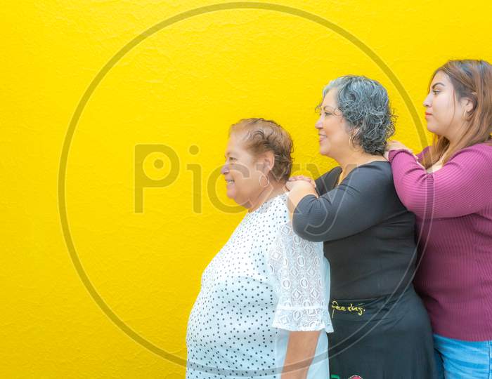 Side View Profiles Of Grandmother, Daughter And Granddaughter Resting On Their Shoulders, Three Generations Of Mexican Women Smiling With Casual Clothes In A Row On A Yellow Background