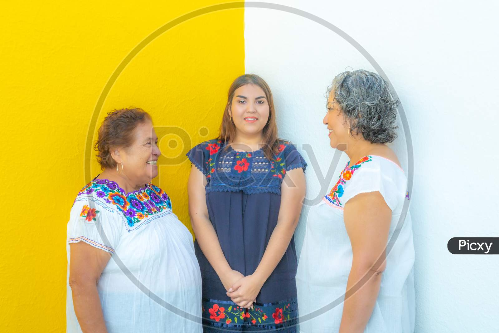 Three Generations Of Mexican Women Smiling With Floral Print Blouses, Mother And Daughter Looking At Each Other And The Granddaughter In The Middle Of Them With A White And Yellow Background