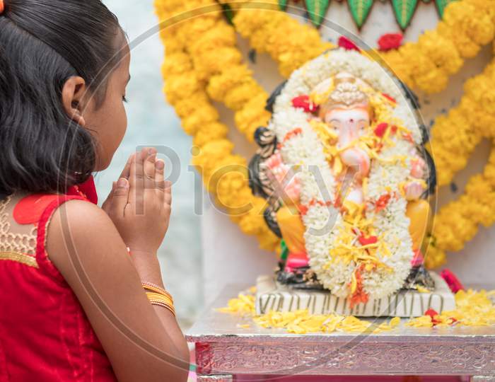 Girl Kid Praying By Closing Eyes And Folding Hands In Front Of Lord Ganesha Idol During Ganesha Or Vinayaka Chaturthi Festival Ceremony At Home - Concept Of Indian Religious Festival Celebrations.