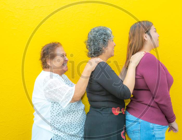Side View Profiles Of Three Generations Of Mexican Women Smiling With Casual Clothes In A Row, Grandmother, Daughter And Granddaughter On A Yellow Background.