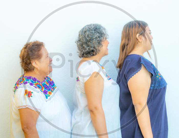 Side View Profiles Of Three Generations Of Mexican Women Smiling With Floral Printed Blouses In A Row, Grandmother, Daughter And Granddaughter On A White Background