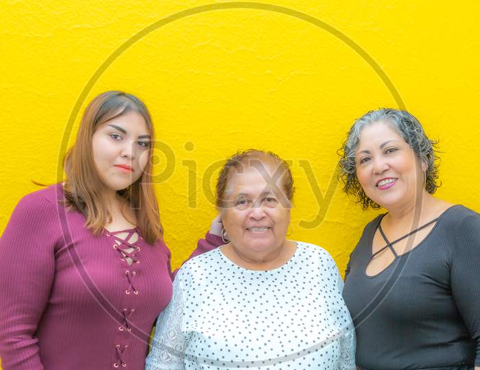 Granddaughter, Grandmother And Daughter Looking At The Camera, Three Generations Of Mexican Women Smiling In Casual Clothes On A Yellow Background