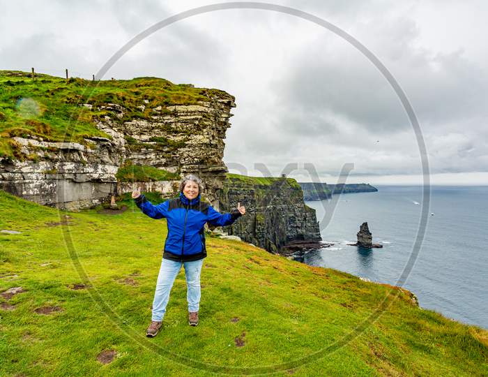 Happy Mexican Mature Woman On The Cliffs Of Moher With The Branaunmore Sea Stack In The Background, Geosites And Geopark, Wild Atlantic Way, Wonderful Spring Cloudy Day In County Clare In Ireland