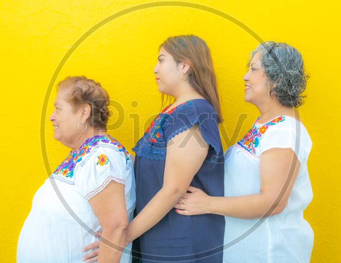 Side View Profiles Of Three Generations Of Mexican Women Smiling With Floral Print Blouses In A Row, Grandmother, Granddaughter And Daughter On A Yellow Background