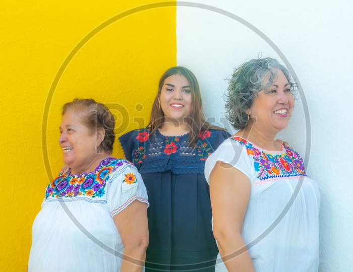 Granddaughter Between Mother And Grandmother Very Cheerful, Three Generations Of Mexican Women Smiling With Floral Print Blouses On A White And Yellow Background