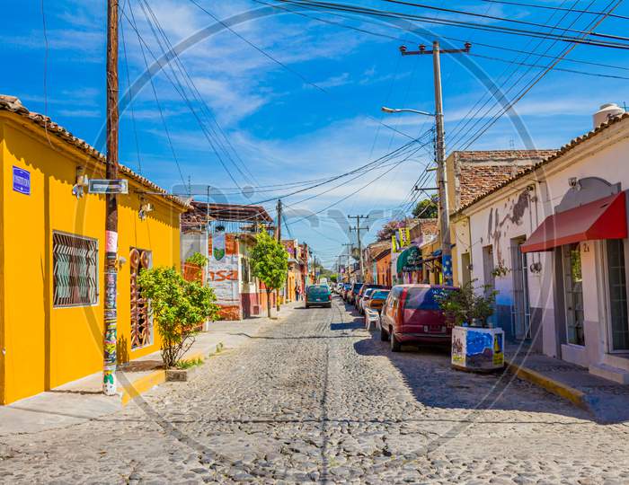 Ajijic, Jalisco / Mexico. February 8Th. 2017. Picturesque Cobblestone Street With Brightly Colored Houses And Shops With Cars Parked Next To A Sidewalk, Electric Poles With Crossover Cables, Sunny Day