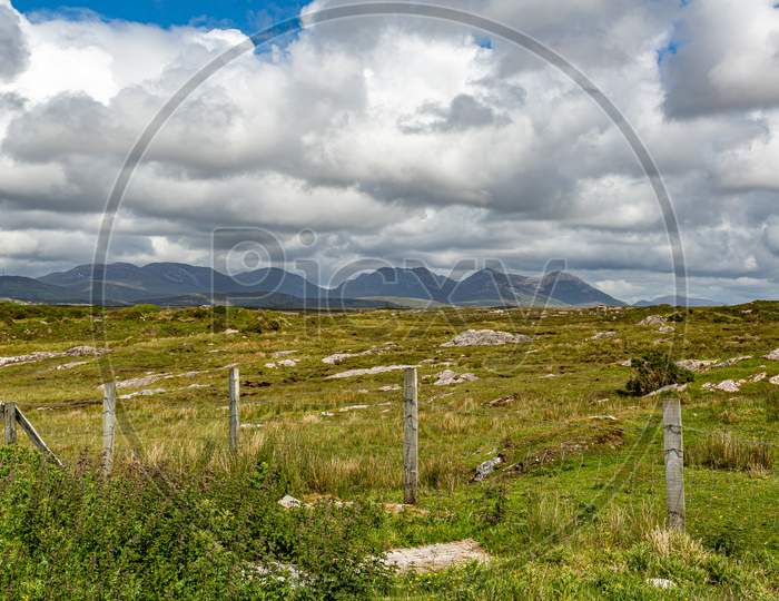 Fences In The Irish Countryside With Green Grass And Limestone Rock Mountains In The Background At Derrigimlagh Bog, Spring Day With Cloudy Sky In County Galway, Ireland