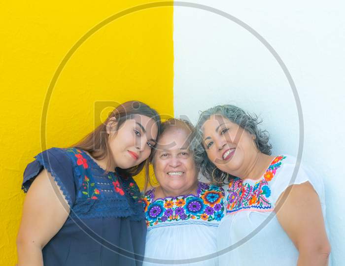Three Generations Of Mexican Women Smiling, Granddaughter And Daughter Leaning On The Mother In Floral Print Blouses Looking At The Camera With A Yellow And White Background.
