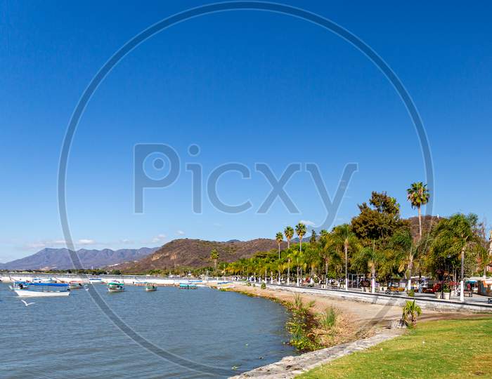 Boardwalk Along The Bay Of Lake Chapala With Motorboats On The Calm Waters, The Pier And Mountains In The Background, Sunny Day With A Clear Blue Sky