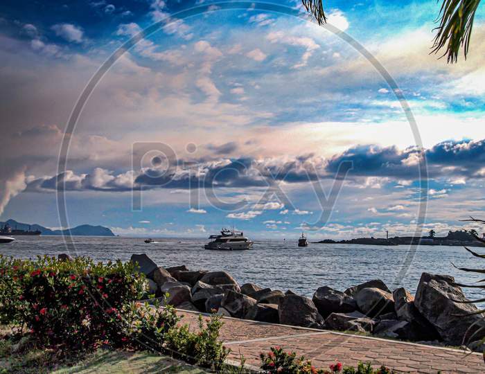 Boardwalk With A Sidewalk, Bushes And Breakwater With The Sea And Boats Sailing In The Background, Tropical Summer Afternoon With A Blue Sky And Abundant Clouds In Manzanillo, Colima Mexico