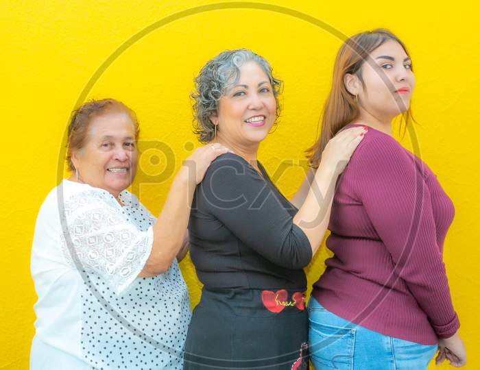 Three Generations Of Smiling Mexican Women Wearing Casual Clothes In A Row Holding Their Shoulders And Looking At The Camera On A Yellow Background