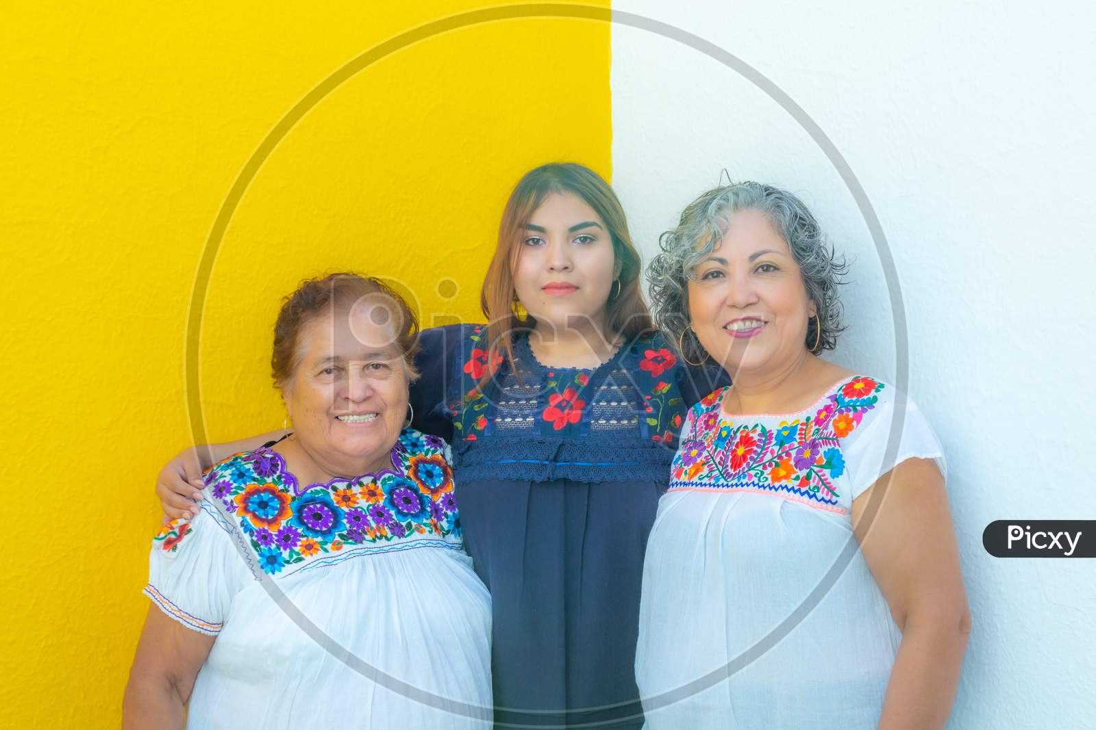 Granddaughter Between Grandmother And Mother Looking At The Camera, Three Generations Of Mexican Women Smiling With Floral Print Blouses On A White And Yellow Background