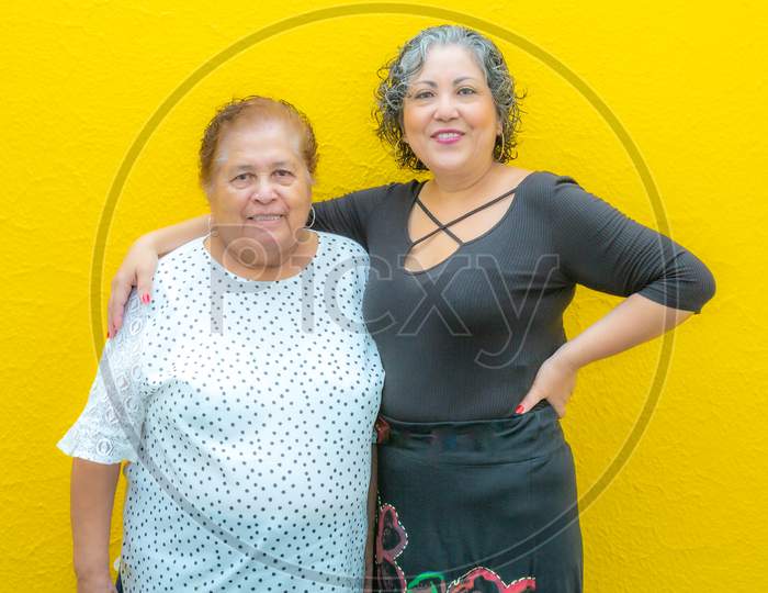 Daughter Hugging Her Mother Very Smiling, Two Mexican Women Wearing Casual Clothes On A Yellow Background