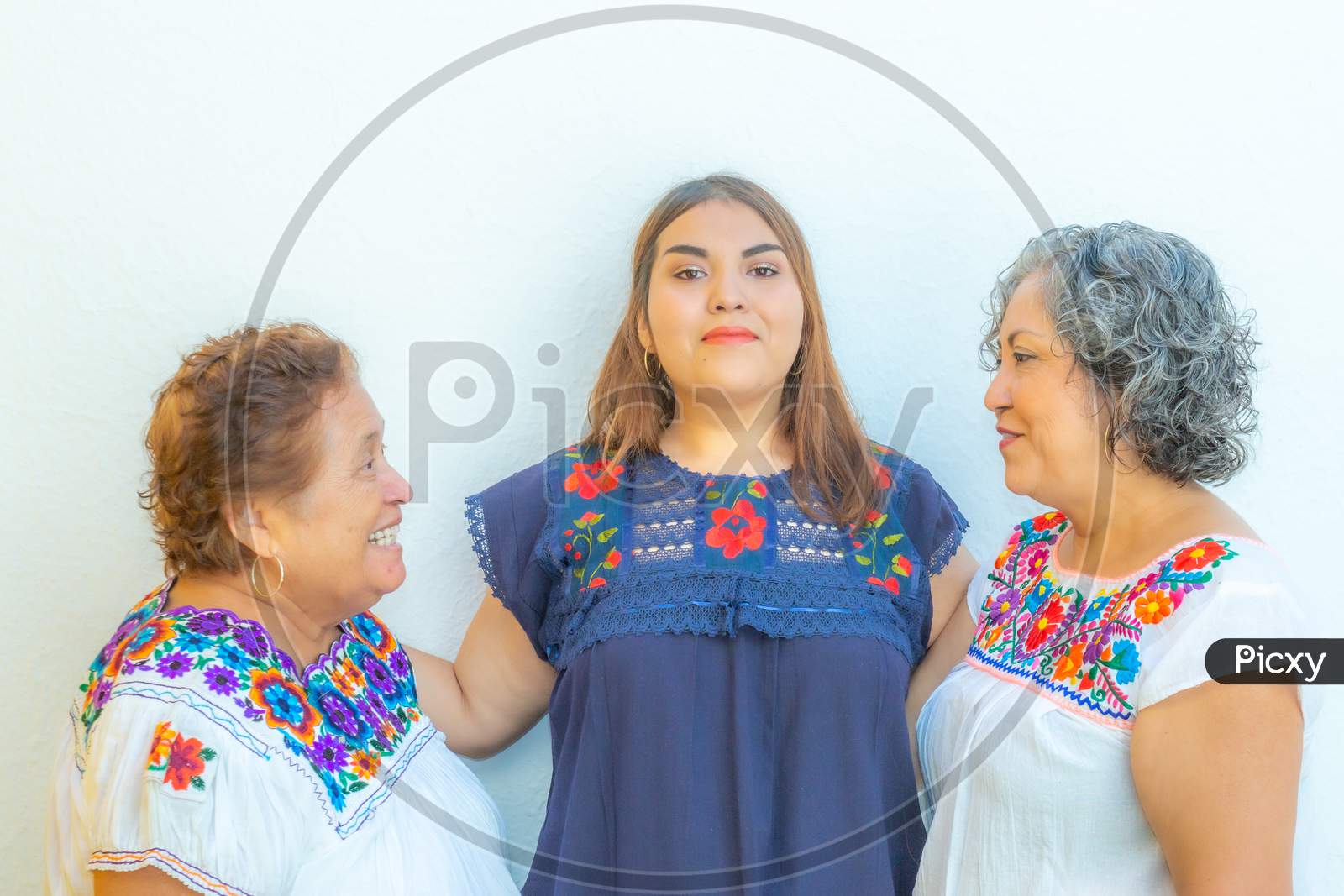 Mother And Daughter Looking At Each Other And  Granddaughter In The Middle Of Them, Three Generations Of Mexican Women Smiling With Floral Print Blouses On A White Background