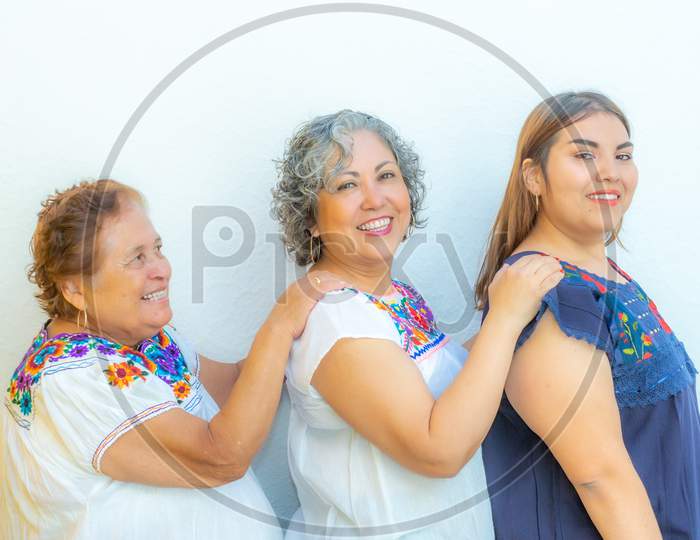 Three Generations Of Smiling Mexican Women With Blouses With Floral Patterns In A Row Holding Their Shoulders Against A White Background