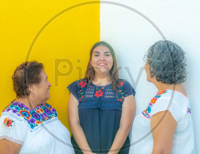 Mother And Grandmother Looking At The Very Cheerful Granddaughter, Three Generations Of Mexican Women Smiling With Floral Print Blouses On A White And Yellow Background