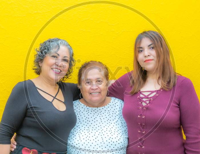 Daughter, Grandmother And Granddaughter Hugging Looking At The Camera, Three Generations Of Mexican Women Smiling In Casual Clothes On A Yellow Background