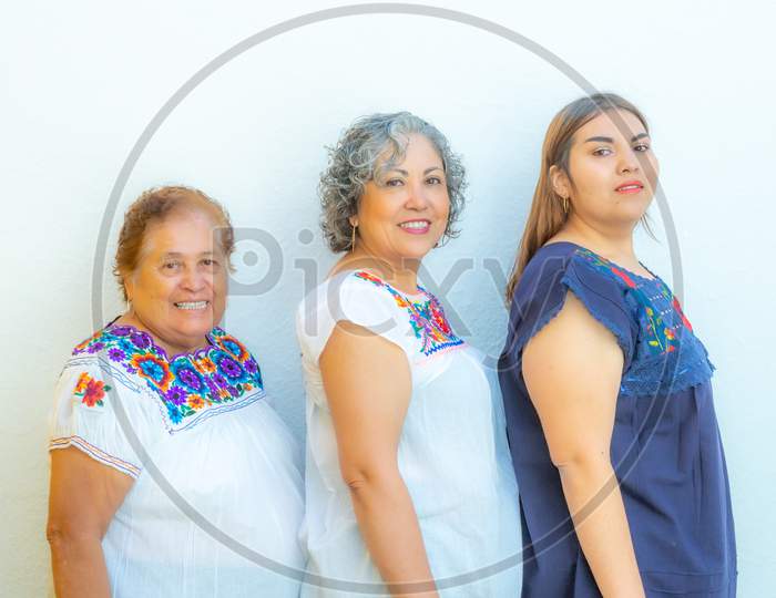 Three Generations Of Smiling Mexican Women With Floral Print Blouses In A Row Looking At The Camera On A White Background