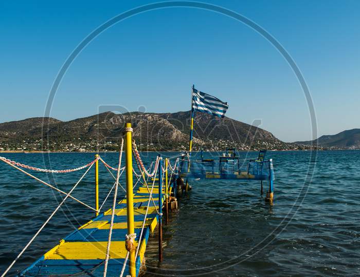 Greek Flag Waves At A Wooden Platform Over The Sea Water Leading To A Table With Three Chairs For Dinner During Summertime Next To The Beach