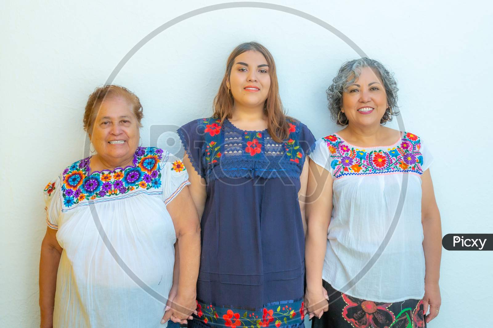 Three Generations Of Latin Mexican Women Smiling, Grandmother, Granddaughter And Daughter With Floral Printed Blouses Holding Hands Looking At The Camera With A White Background