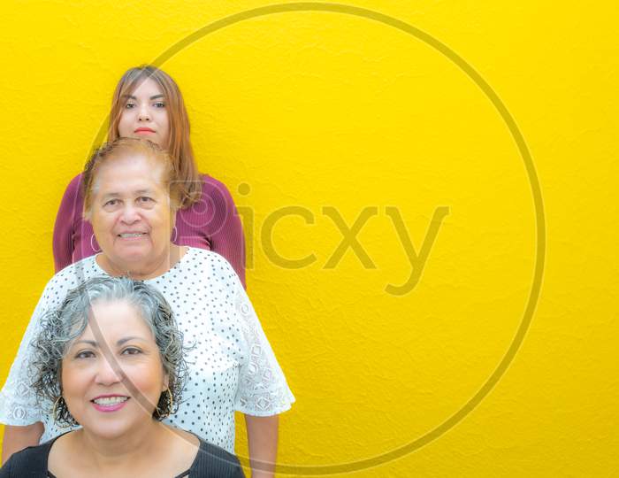 Three Generations Of Latin Mexican Women Smiling In Line, Daughter, Grandmother And Granddaughter Wearing Casual Clothes Looking At The Camera With A Yellow Background, Space For Text