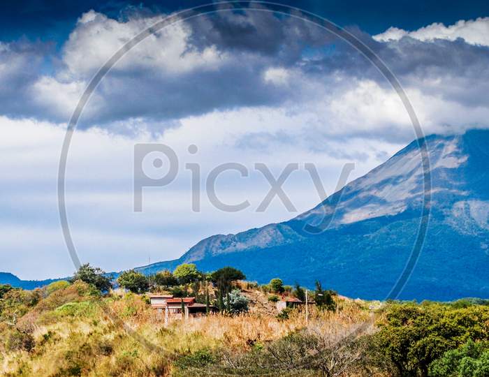 Mexican Landscape With A House On A Hill With The Colima Volcano With A Fumarole In The Background, Sunny Day With A Blue Sky And Abundant Clouds In The State Of Jalisco, Mexico