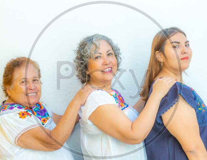 Three Generations Of Smiling Mexican Women With Floral Print Blouses In A Row Holding Their Shoulders And Looking At The Camera On A White Background