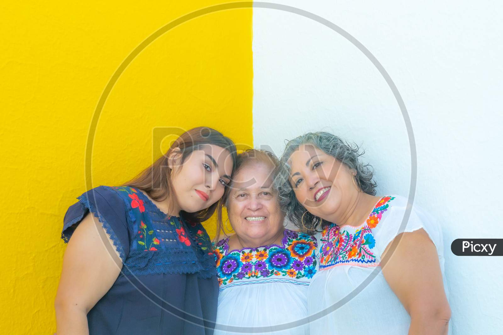 Three Generations Of Mexican Women Smiling, Granddaughter And Daughter Leaning On The Mother In Floral Print Blouses Looking At The Camera With A Yellow And White Background.