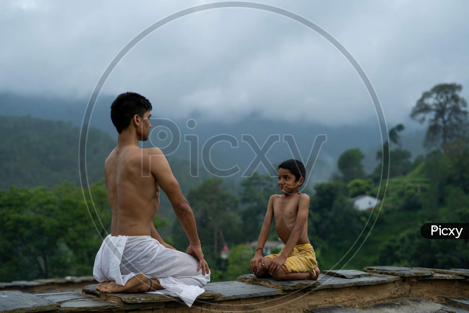 A Young Handsome Boy Doing Yoga With A Kid On The Roof Of A House Situated In The Middle Of Mountain Range. Yoga And Fitness.