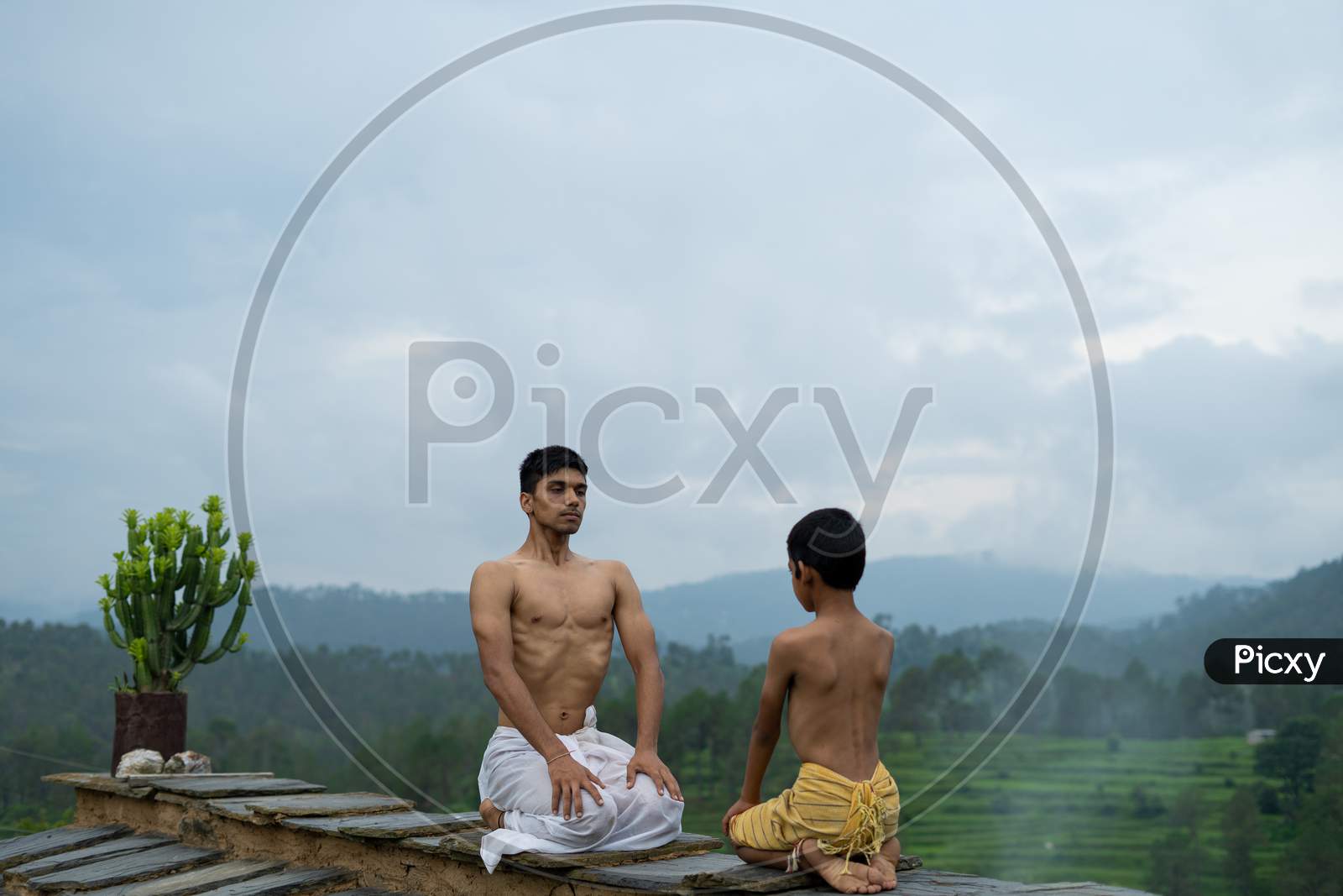 A Young Handsome Boy Doing Yoga With A Kid On The Roof Of A House Situated In The Middle Of Mountain Range. Yoga And Fitness.