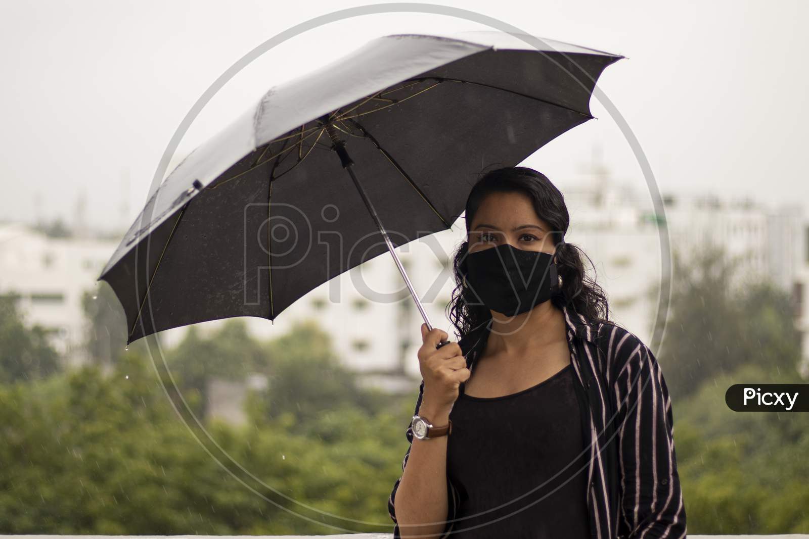 Indian Woman With Umbrella In The Rain. Looking At Camera.Covid-19 Pandemic Coronavirus A Young Indian Woman In A Protective Black Mask For The Spread Of The Sars-Cov-2 Disease Virus.