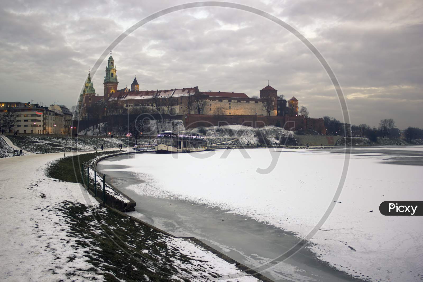 Krakow, Poland - January 09, 2016: Wide Angle View Of Famous Wawel Castle Covered With Snow Next To Vistual River Against Dramatic Clouds