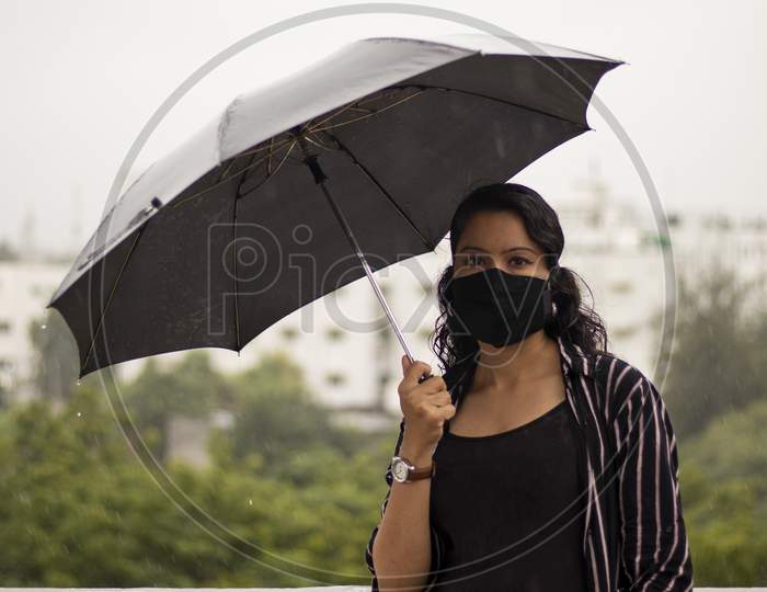 Indian Woman With Umbrella In The Rain. Looking At Camera.Covid-19 Pandemic Coronavirus A Young Indian Woman In A Protective Black Mask For The Spread Of The Sars-Cov-2 Disease Virus.