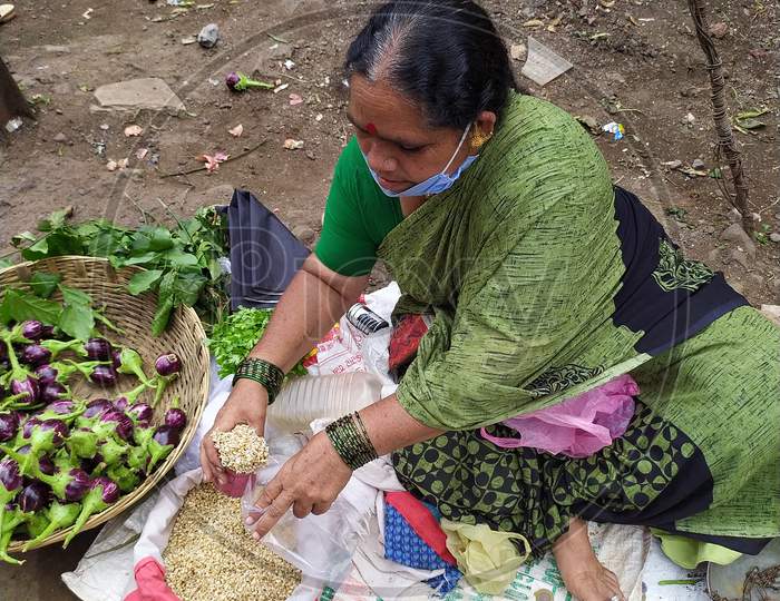 Aged lady wear face mask and selling vegetables in market yard. Measuring moth bean legumes with use of measure utensil..