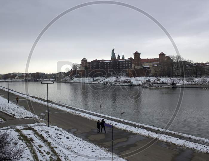 Krakow, Poland - January 29, 2015: Wide Angle View Of Famous Wawel Castle Covered With Snow Next To Vistual River Against Cloudy Sky
