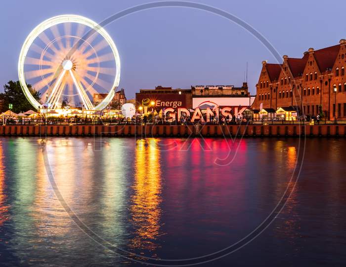Gdansk, North Poland: Wide Angle Night View Of Ferris Wheel In Motion And City Center With Gdansk Sign And Reflection On Mouth Of The Motława River Near Baltic Sea