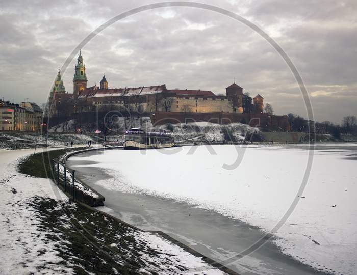 Krakow, Poland - January 09, 2016: Wide Angle View Of Famous Wawel Castle Covered With Snow Next To Vistual River Against Dramatic Clouds