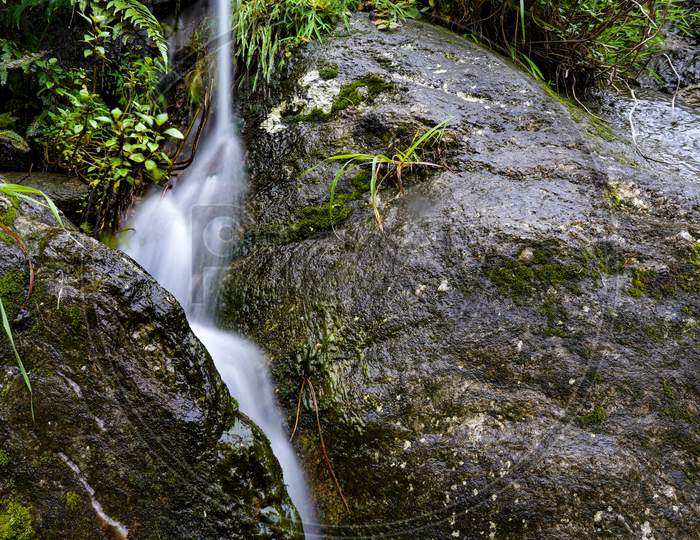 Slow Shutter Of Water Peeping Through Rocks With Natural Vegetation Around It.