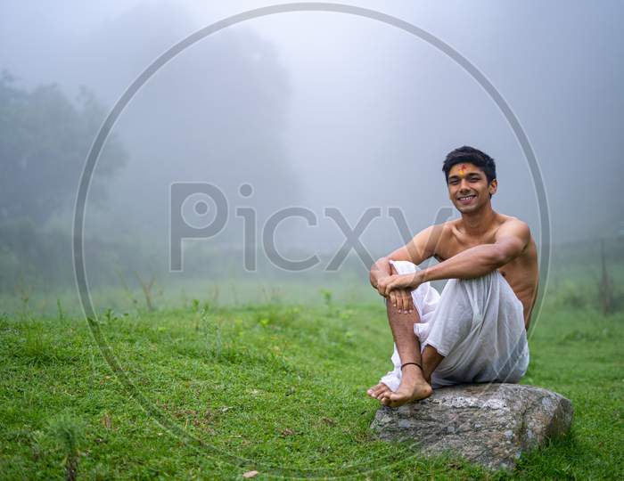 Young Man Wearing A Dhoti, Sitting On A Rock, Open In The Green Field In A Foggy Morning.