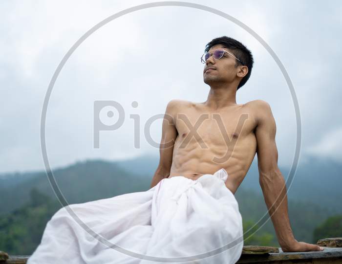 Young Shredded Man Sitting Wearing A Dhoti And Glasses With Blurred Mountains In The Background.